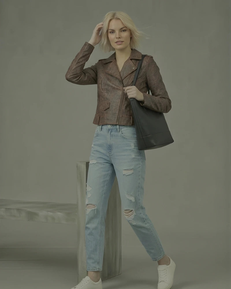 Woman wearing a brown leather jacket and light blue jeans, holding a black handbag against a gray background.