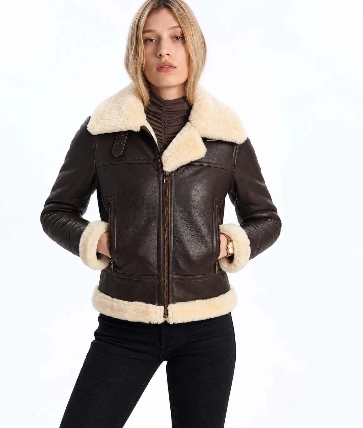 A woman wearing a dark brown leather aviator-style jacket with a thick cream-colored shearling collar, cuffs, and hem, paired with a dark brown turtleneck and black pants, against a plain white background.